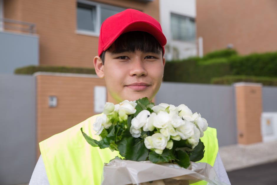 A young male delivery person with light skin, wearing a red cap and a high-visibility yellow vest, is holding a bouquet of fresh white roses wrapped in beige paper with green leaves visible. He is out