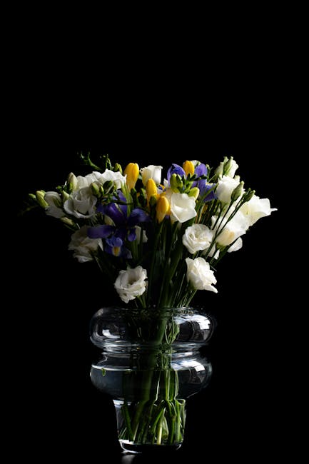 A vibrant flower arrangement in a clear glass vase featuring fresh white roses, deep purple irises, and bright yellow freesias, with green stems and leaves visible through the transparent glass. The b