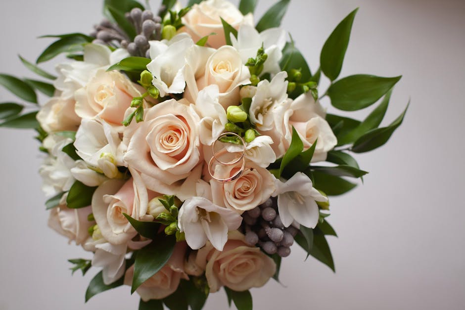 A close-up view of a bouquet featuring pale peach roses, white freesias, and small green buds, accented with dark green foliage and sprigs of grayish-purple berries. The flowers are fresh, with soft, 