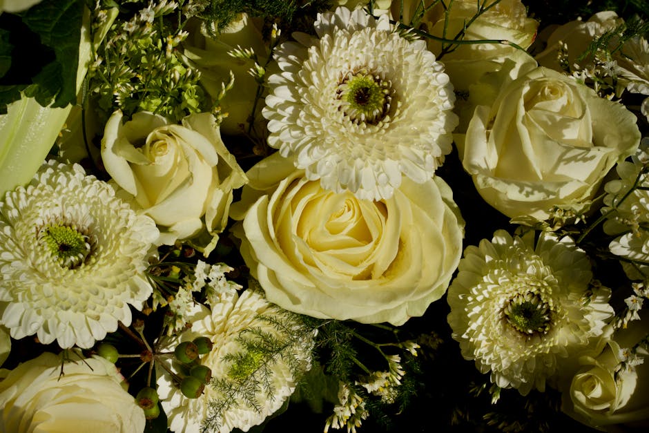 A close-up arrangement of fresh white flowers, including large roses with softly curled petals, complemented by white chrysanthemums with densely packed petals and green centers, all set against dark 