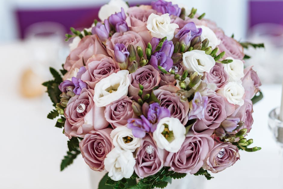 A close-up view of a floral bouquet arranged with soft pink roses, white lisianthus, and lavender-purple freesia, accented with small green buds and lush foliage. The flowers are fresh, with delicate 