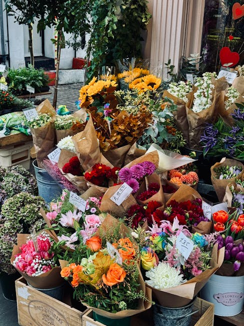 An outdoor floral display featuring an array of fresh flower bouquets arranged in brown craft paper wrapping and metal buckets. The selection includes sunflowers, pink lilies, red and purple carnation